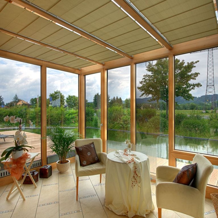 Wintergarten von innen mit Holzrahmen, Stoffbeschattung unter dem Glasdach, Sesseln und Blick auf eine Wiese