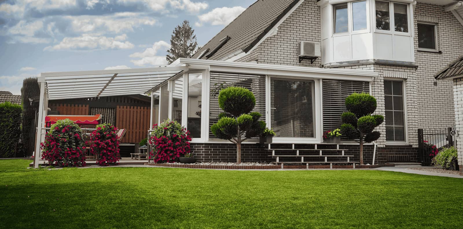 Weißer Wintergarten mit angrenzender Terrassenüberdachung und Raffstore in einem gepflegten Garten