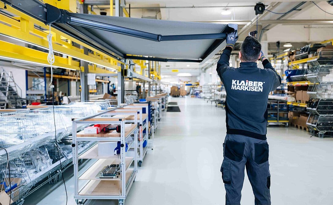 Produktionshalle mit Mitarbeiter in Arbeitskleidung, der eine ausgefahrene Kassettenmarkise mit Werkzeug montiert