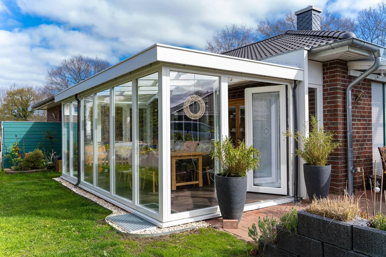 Weißer Wohnwintergarten mit Flachdach und raumhoher Glasfront an einem Klinkerhaus (Schennjesse)